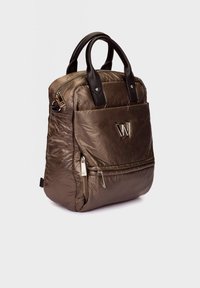 Sac à dos matelassé brun avec poignées noires, poche zippée à l'avant et logo métallique "W" sur un fond clair uni.