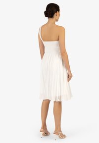Bílá skládací šaty se smaragdovým živůtkem, jedním ramínkem a splývavou sukní. Doplněné světlými sandály na podpatku.