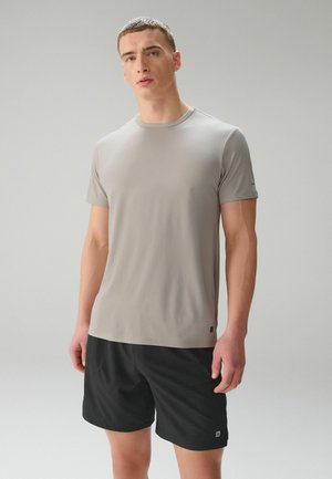 Ung mand stående med front mod kameraet iført en lys grå kortærmet skjorte og sorte shorts mod en ensfarvet grå baggrund.