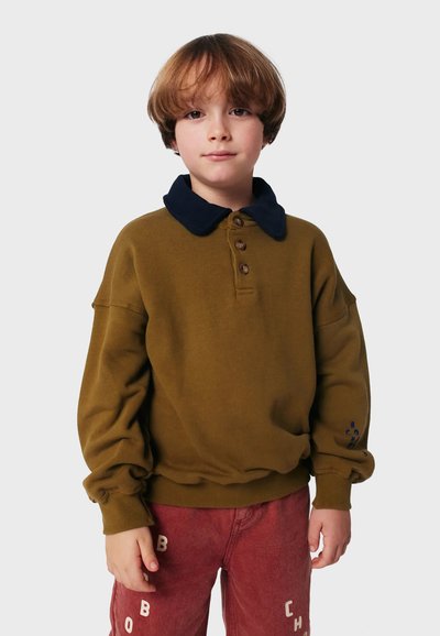 Sweatshirt vert olive avec col marine et trois boutons marron, coupe ample, poignets côtelés, associé à un pantalon rouge avec des lettres blanches.