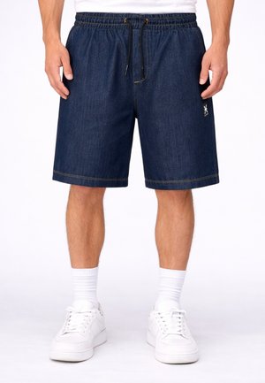 Uomo che indossa pantaloncini di jeans blu scuro con vita elasticizzata e coulisse, calzini bianchi e scarpe da ginnastica bianche, in piedi su sfondo bianco uniforme.