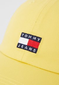 Căciulă galbenă din bumbac, cu un patch dreptunghiular cusut pe care scrie "TOMMY JEANS" și un logo în culorile roșu, alb și albastru.