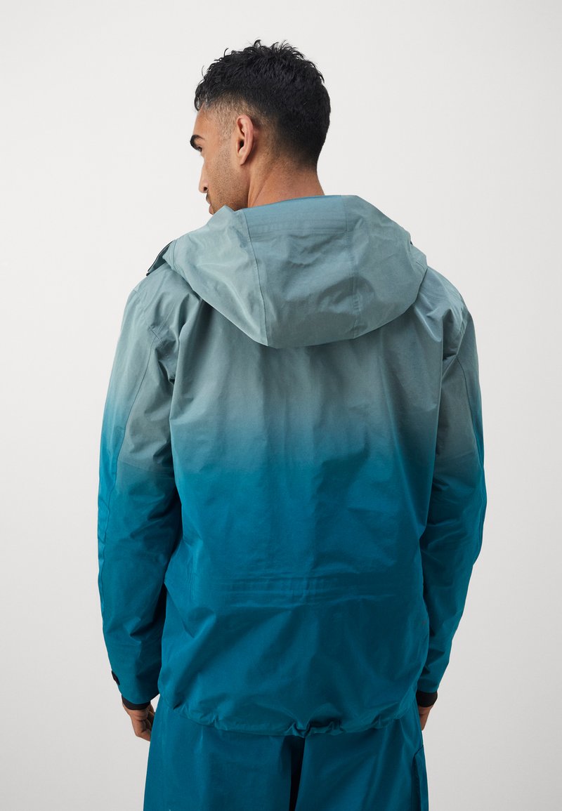 Let jakke i gradient nuancer af teal og azur, med hætte, justerbar kant og tekstureret stof for åndbarhed.