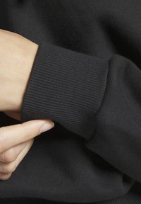 Ruka upravuje žebrovaný manžet černého dlouhého rukávu oděvu, ukazuje texturu látky a detail rukávu.