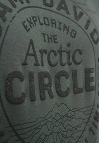 Dark green fabric featuring a textured black graphic that reads "Exploring the Arctic Circle" with line art of mountains below.