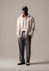 Light beige oversized shirt over a white t-shirt, gray trousers, black and white shoes, and a dark cap. Smooth fabric, relaxed fit.