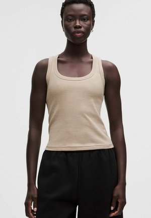 Giovane persona dalla pelle scura che indossa un top beige senza maniche e pantaloni neri, in piedi dritta contro uno sfondo grigio chiaro.