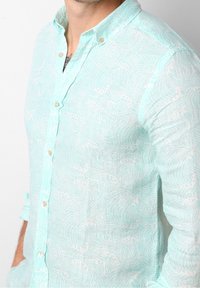 Chemise bleu clair en lin à boutons, présentant un motif de vagues, un col et des manches retroussées avec des boutons beige.