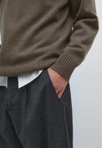 Bruine gebreide trui met geribbelde mouwen, gedragen over een wit gestreept shirt, gecombineerd met donkergrijze gestructureerde broek. Hand in de zak.