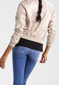 Světle béžová saténová bomber bunda s žebrovanými manžetami, nošená přes černý top, v kombinaci s přiléhavými modrými džínami. Design se vyznačuje hladkou texturou.