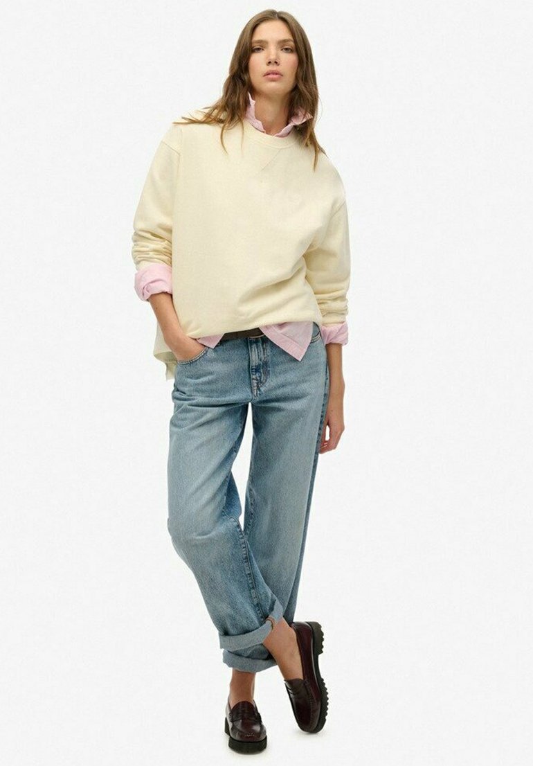 Sweatshirt crème surdimensionné superposé à une chemise rose clair, associé à un jean bleu roulé et des mocassins foncés avec une semelle texturée.