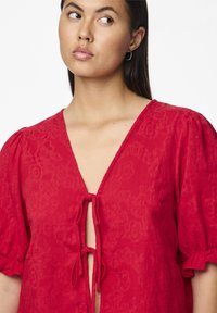 Blusa rossa con tessuto testurizzato, scollo a V profondo e dettaglio con fiocco sul petto. Maniche corte a palloncino per aggiungere volume e stile.
