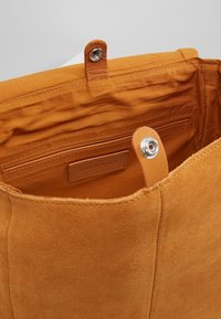 Bolso de ante color canela con interior forrado de tela suave, que incluye un bolsillo con cremallera y cierre a presión. Etiqueta de la marca visible en cuero.