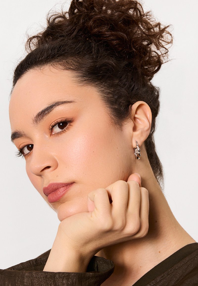 Srebrna kolczyk w kształcie obręczy z liściastym wzorem; modelka ma kręcone włosy i opiera brodę na dłoni, ubrana w strukturalny ciemny top.