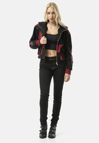 Zwart en bordeaux cropped jack met ritssluiting, voorzien van een capuchon en rode accenten, gecombineerd met zwarte skinny jeans en enkellaarsjes met gesp.