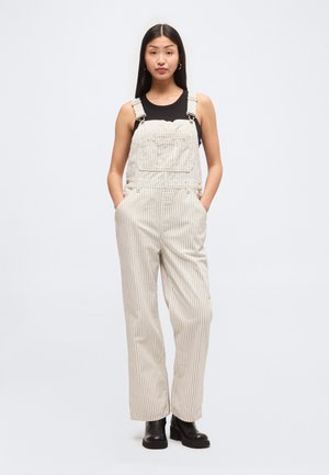 Γυναίκα στέκεται με τα χέρια στις τσέπες, φοράει κρεμ ριγέ παντελονόφορεμα, μαύρο αμάνικο τοπ, κρίκους σκουλαρίκια και μαύρα μποτάκια μέχρι τον αστράγαλο.