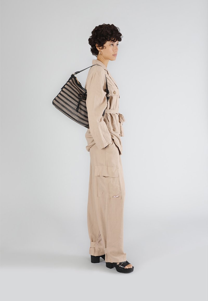 Beige jumpsuit med bälte i midjan, stora cargofickor och avslappnad passform. Svart handväska med vävt mönster, bärs på axeln.