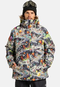 Mand iført en farverig, mønstret vinterjakke med rummet og aliens illustrationer, sort beanie og goggles på panden, hænderne i lommerne.