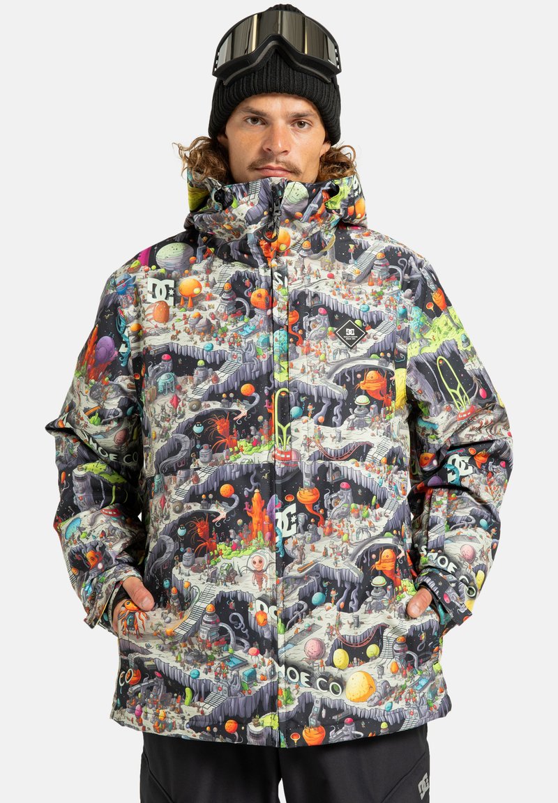 Mand iført en farverig, mønstret vinterjakke med rummet og aliens illustrationer, sort beanie og goggles på panden, hænderne i lommerne.