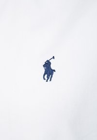 Weißes Poloshirt mit einem in Marineblau gestickten Logo eines Pferdes mit Reiter. Der Stoff ist glatt und weist eine minimale Textur auf.