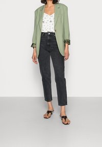 Donna che indossa un blazer verde salvia sopra una blusa bianca a fiori, con jeans neri a vita alta e sandali con tacco marrone scuro.