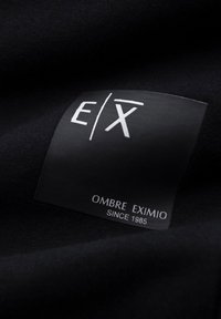 Černá látka s oděvním štítkem s bílým textem "E/X OMBRE EXIMIO SINCE 1985" na matně černém pozadí.