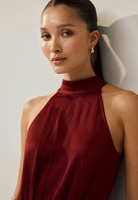Vinrød halterneck-kjole med en tekstureret overflade, der har høj krave og ærmeløst design. Glat stof med en let glans.