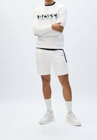 Bílá mikina s černo-tyrkysovým logem, spárovaná s bílými šortkami s bočními kapsami a tyrkysovými detaily, nošena s odpovídajícími teniskami a ponožkami.