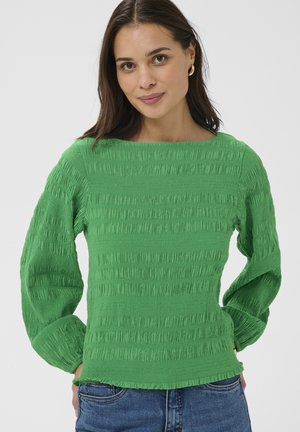 Femme aux longs cheveux bruns portant des boucles d'oreilles en or, un haut vert texturé à manches longues et un jean bleu sur un fond uni.