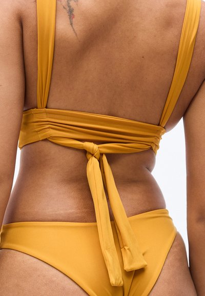 Μπικίνι σε μουσταρδί κίτρινο με δέσιμο στο πίσω μέρος και φαρδιές τιράντες. Λείο ύφασμα, μίνιμαλ σχεδιασμός, που αναδεικνύει μια εφαρμοστή σιλουέτα με έναν δεμένο κόμπο.