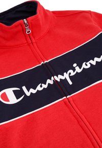 Giacca zip-up rossa con colletto alto, caratterizzata da una striscia orizzontale blu navy e dal logo bianco "Champion". Tessuto morbido con finitura testurizzata.