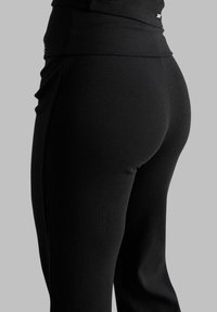 Gros plan d'une personne vêtue d'un pantalon noir côtelé ajusté et d'un haut noir assorti sur un fond gris uni.