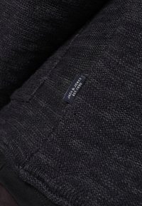 Ciemny sweter dziany o teksturowanej tkaninie, z małą niebieską metką przyszytą z boku, na której widnieje napis "JACK & JONES EST. 1990."