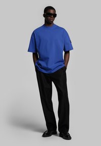 T-shirt bleu oversize à manches courtes, associé à un pantalon noir ajusté et des chaussures noires. Modèle portant des lunettes de soleil sombres, fond neutre.