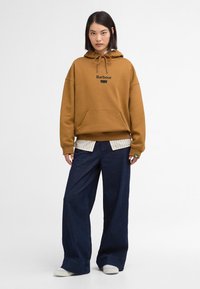 Καφέ φούτερ με μπροστινή τσέπη και κουκούλα με κορδόνι, που φέρει το μαύρο λογότυπο "Barbour". Φοριέται πάνω από μια ριγέ μπλούζα και συνδυάζεται με φαρδιά μπλε τζιν.