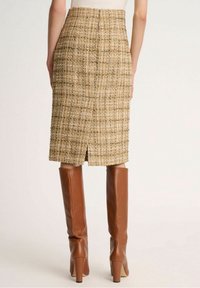 Jupe crayon en tweed beige avec un motif texturé et une fente arrière, associée à des bottes en cuir marron jusqu'aux genoux.