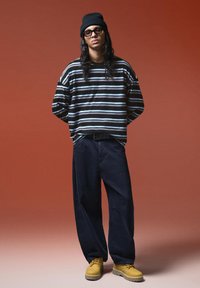 Fekete csíkos hosszú ujjú ing fehér és kék csíkokkal, sötét széles lábú nadrággal és sárga fűzős csizmával párosítva. Fekete sapka van rajta.