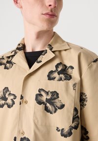 Beige ing fekete hibiszkusz virágmintával, rövid ujjú, gallérral és elülső zsebbel. Könnyű anyagból készült, sima textúrával.