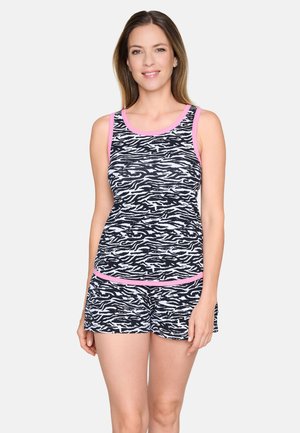 Femme portant un débardeur à imprimé zèbre noir et blanc et un short assorti avec une bordure rose, debout et souriante devant un fond uni.