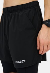 Pantalones cortos de running negros con una cinturilla elástica, bolsillos laterales y un logo "cep" en blanco. El usuario lleva un reloj deportivo en la muñeca.