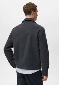 Giacca grigio scuro con colletto a revers, realizzata in tessuto strutturato. Caratterizzata da maniche lunghe e polsini con bottoni, indossata sopra una camicia a righe bianche.