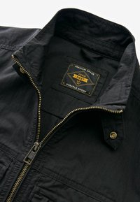 Veste noire avec fermeture éclair en laiton partiellement zippée, col élevé, et étiquette à l'intérieur indiquant "Premium Grade 1982 Next Durable Goods."