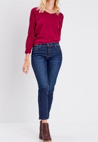 Magenta Langarmshirt mit Spitzenakzenten an den Ärmeln, kombiniert mit dunkelblauen Slim-Fit-Jeans und braunen Stiefeletten. Sauberes, modernes Design.