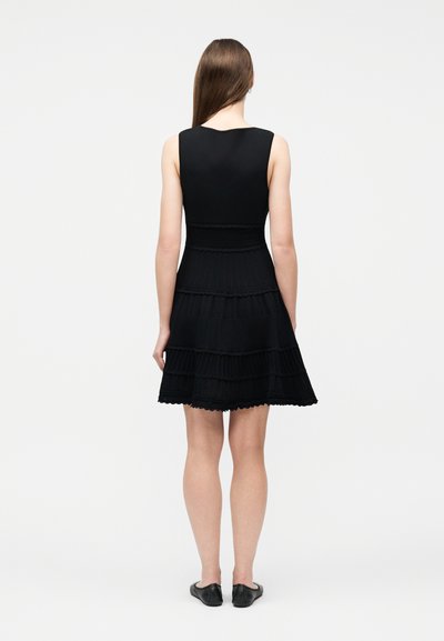 Femme debout de dos, portant une robe noire sans manches à volants superposés et des chaussures plates noires, sur un fond blanc uni.