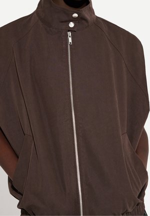 Dark brown zip-up jacket with high collar and snap buttons, hands in pockets, shown from chest to hips.