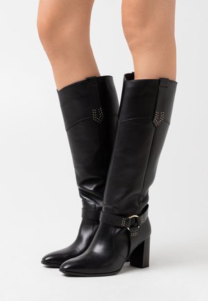 Černé kožené boty ke kolenům s blokovým podpatkem, špičatou špičkou, ozdobnými cvočky a páskem s kovovým kroužkem kolem kotníku.