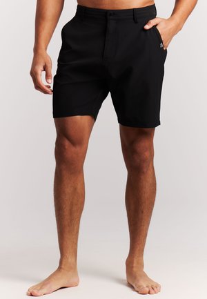 Man die zwarte kniebroek draagt met één hand in zijn zak, staat op blote voeten tegen een effen lichte achtergrond.