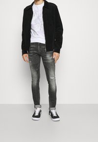 Chaqueta de pana negra sobre una camiseta blanca con un diseño gráfico de rostros abstractos; combinada con unos jeans ajustados y desgastados en gris y zapatillas negras.