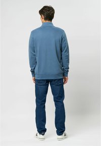 Blue fleece sweatshirt with a collar, paired with dark blue jeans and white sneakers, viewed from the back against a light background.