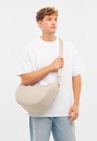 Beige válltáska sima felülettel, íves formával és állítható pánttal, amit a vállon tartanak egy egyszínű háttér előtt.